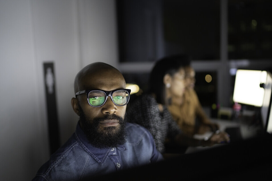 Black male professional looking forward with 2 people seated behind him, looking at computer screens..