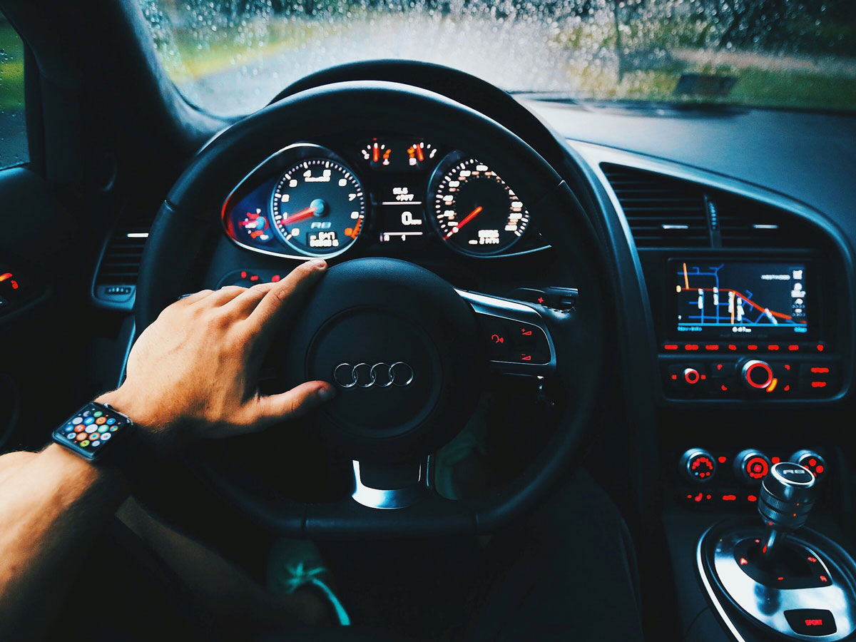 Close-up of a man’s left hand on car steering wheel with view of dashboard and center console.