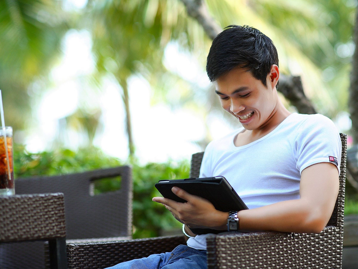 Smiling man sitting outdoors, looking down at a tablet device that he’s holding.