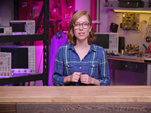 Woman in blue plaid shirt standing behind a counter with tools and a motherboard hanging on the back walls. decoding=